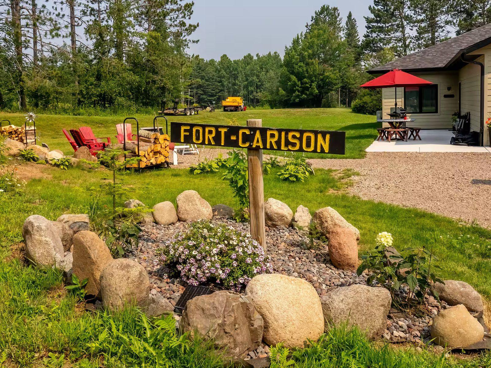 Fort Carlson from the driveway: a hand-painted sign in a fieldstone flower bed, red Adirondack chairs, stacked firewood, and a single-level house with a red patio umbrella.